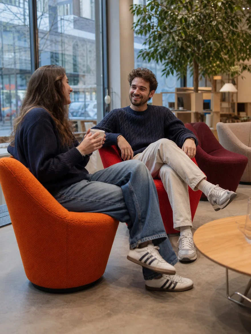 Drehbare Loungesessel in Orange- und Rottönen im Einsatz | Moderne Polsterei Drehbare Loungesessel in Orange- und Rottönen in der Lounge eines Design-Showrooms, zwei Freunde lachen: Moderne Polsterei